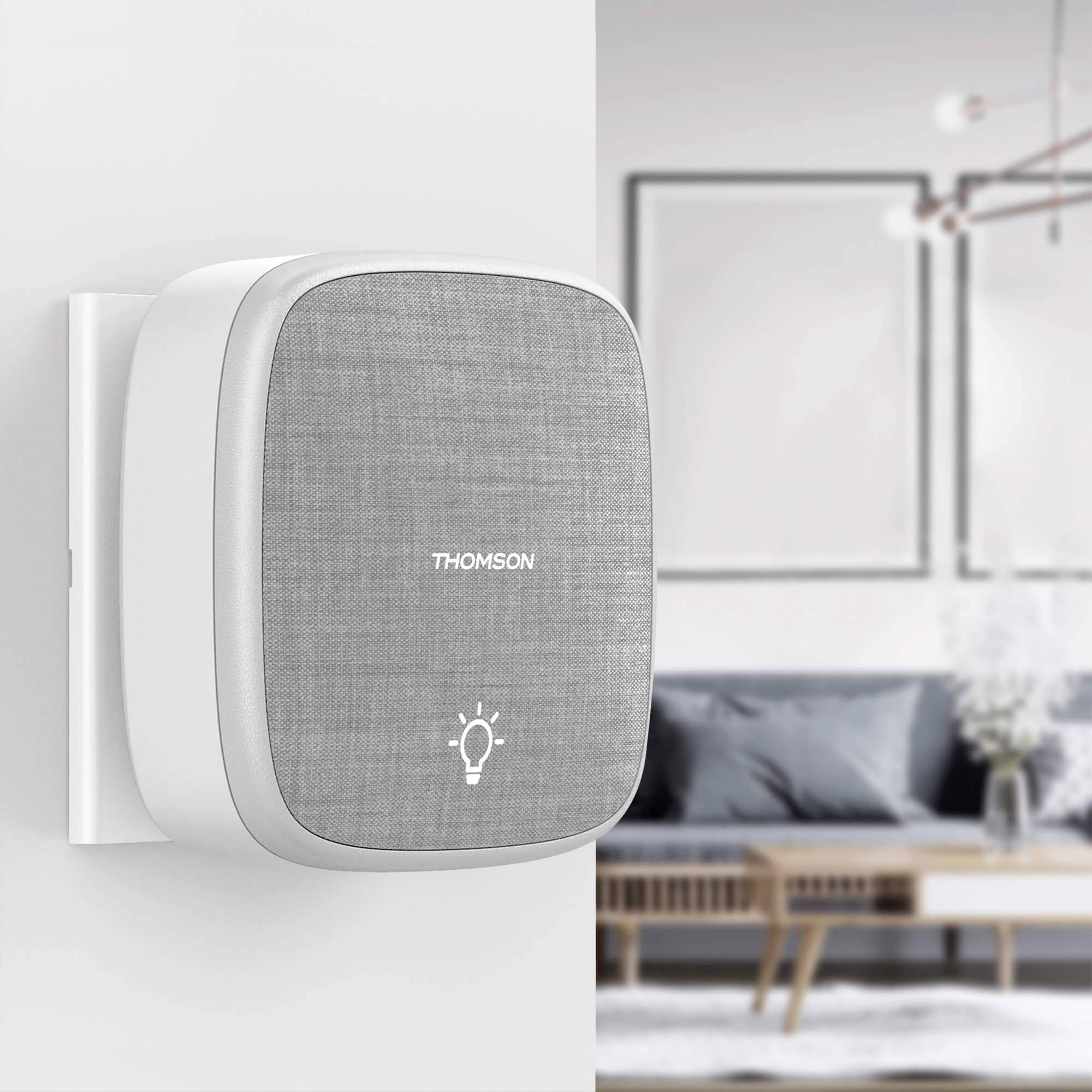 A smart speaker by Thomson is hanging on the wall in a modern living room. A sofa and picture frames can be seen in the background.