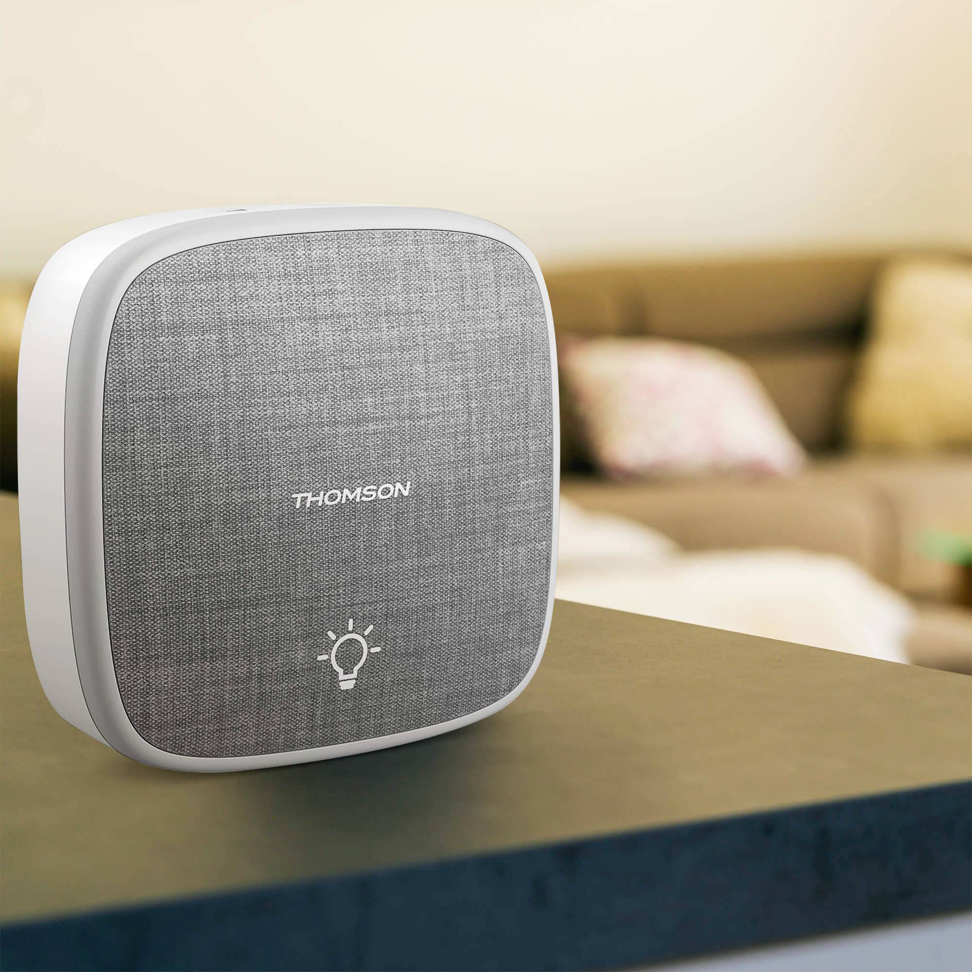 A grey, square speaker with the 'Thomson' logo sits on a table. A blurry living room is visible in the background.