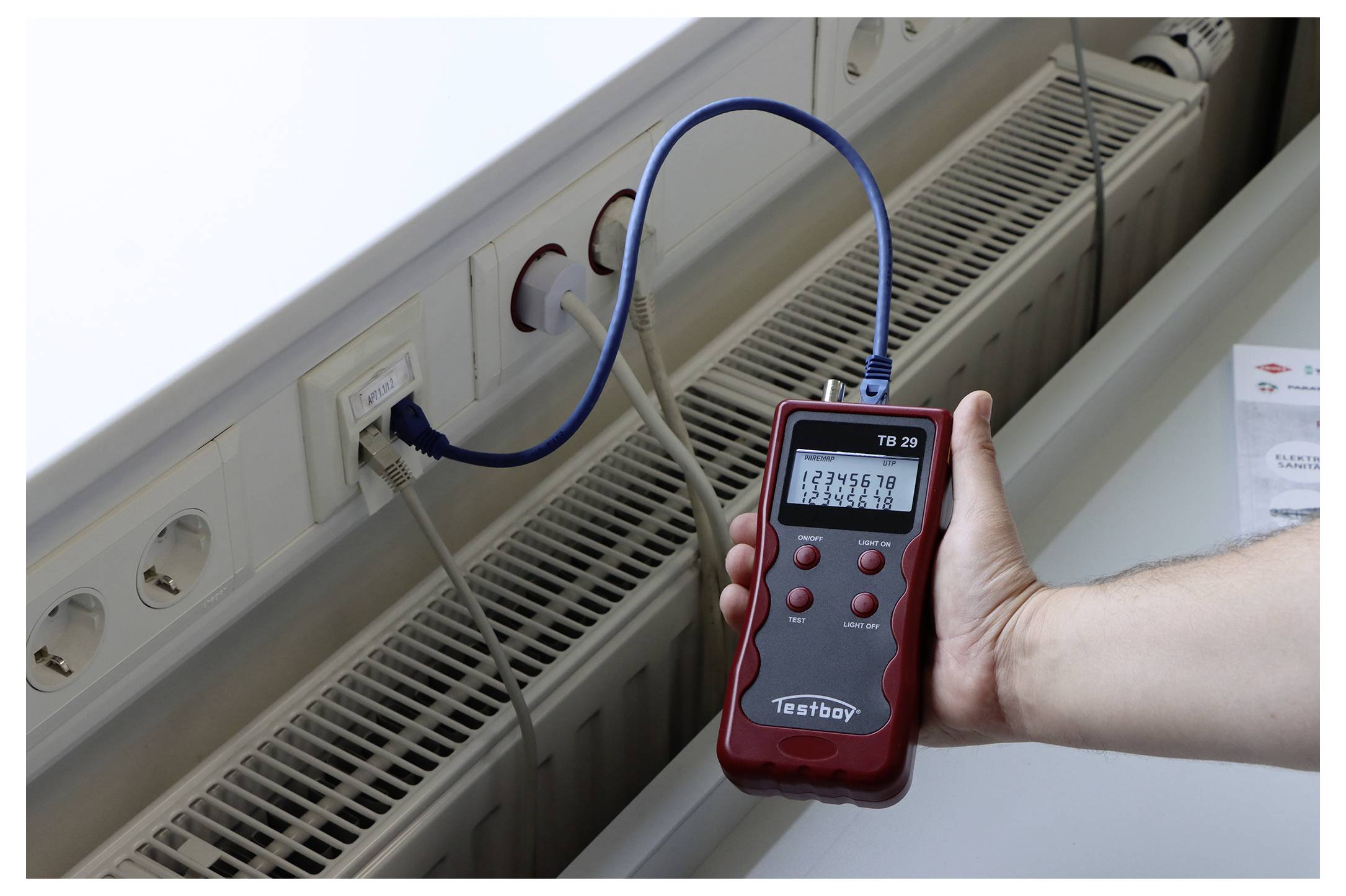 'A hand holds a red network tester displaying numbers, connected to a network port with a blue cable, indicating a test in progress.'