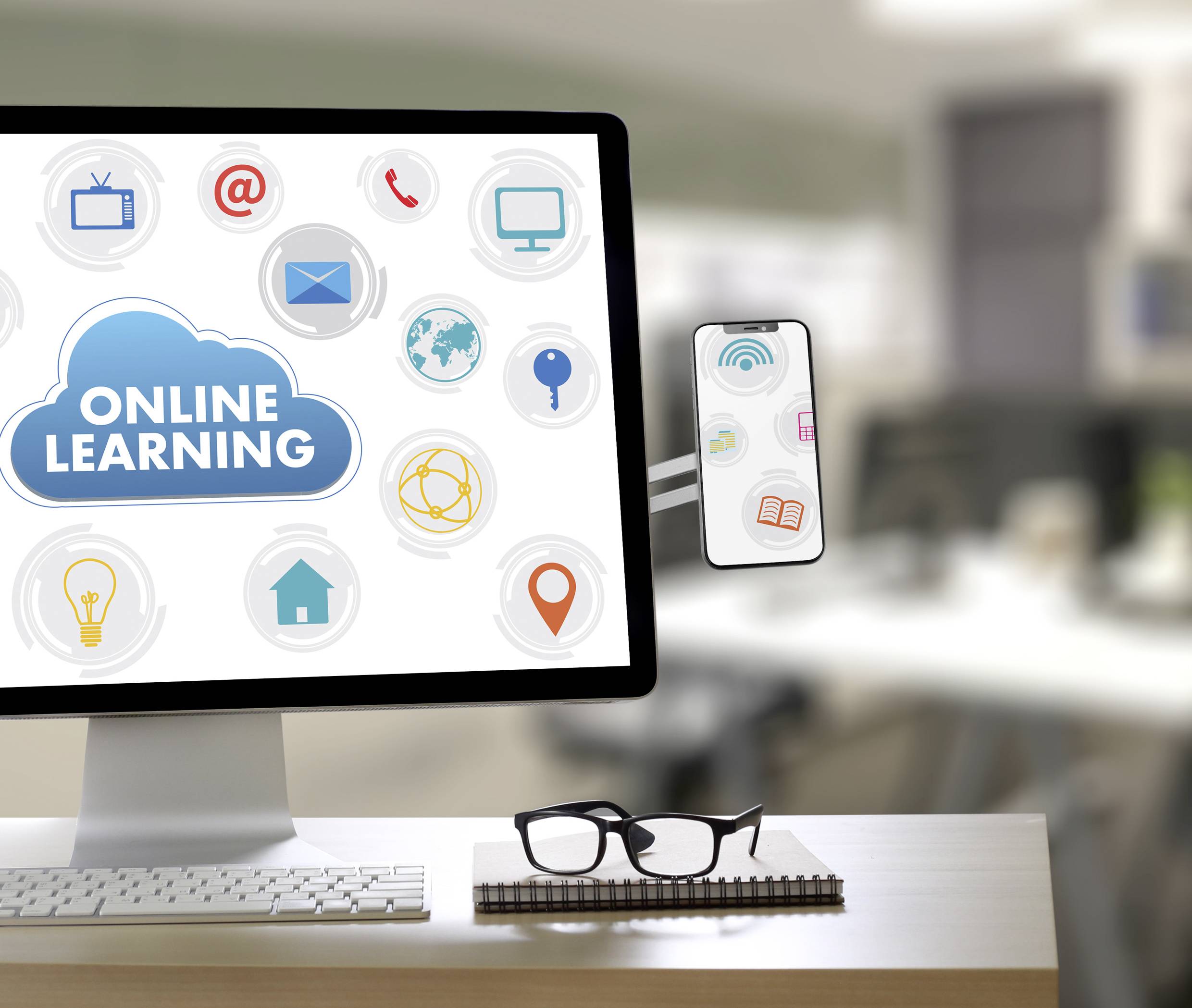 On a desk, there is a computer screen displaying 'Online Learning' text and symbols, alongside a pair of glasses. The background is blurred.