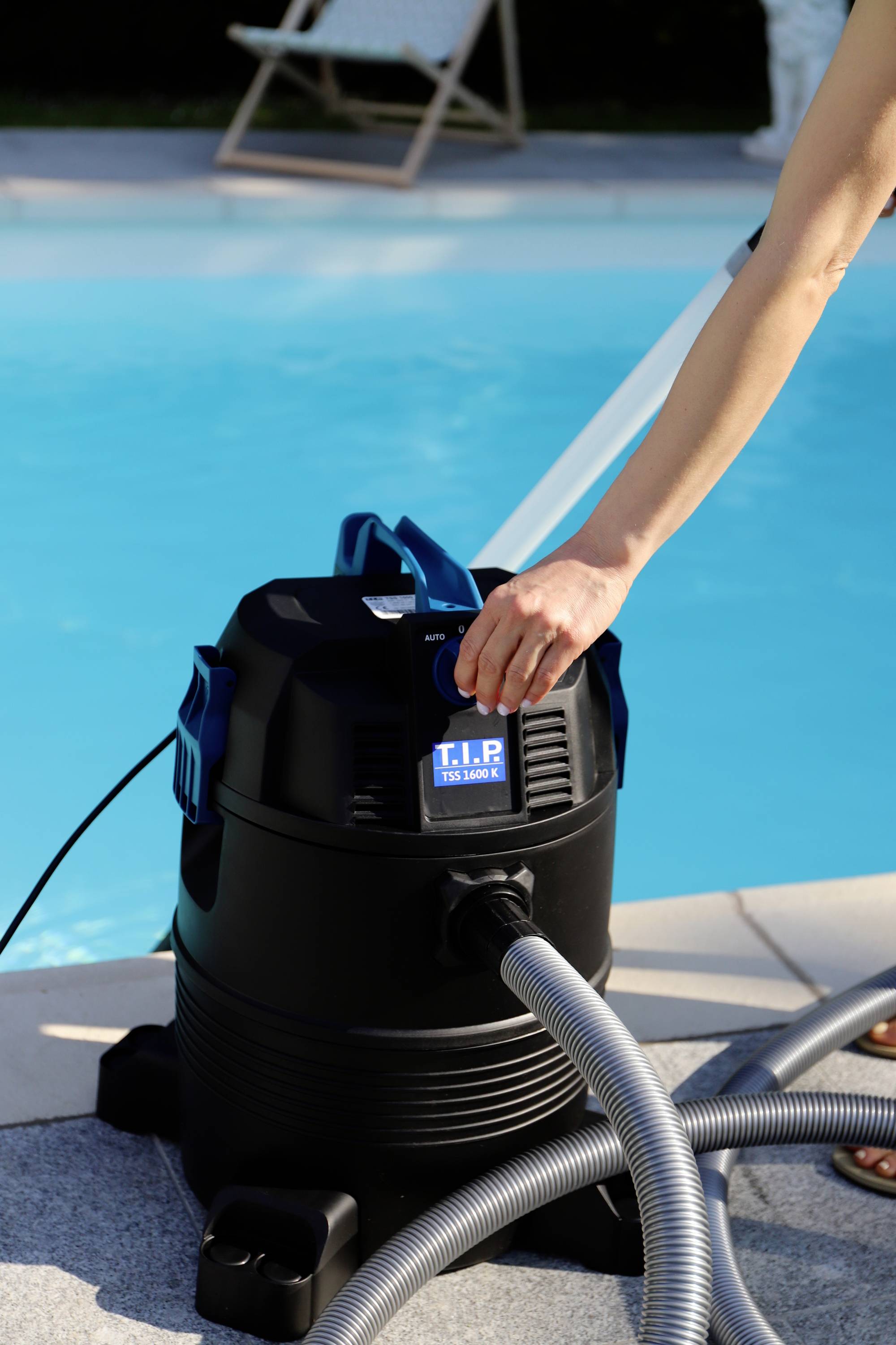 A person is cleaning a pool with a black vacuum cleaner. The suction pipe and hoses are connected to the device. Summery surroundings.