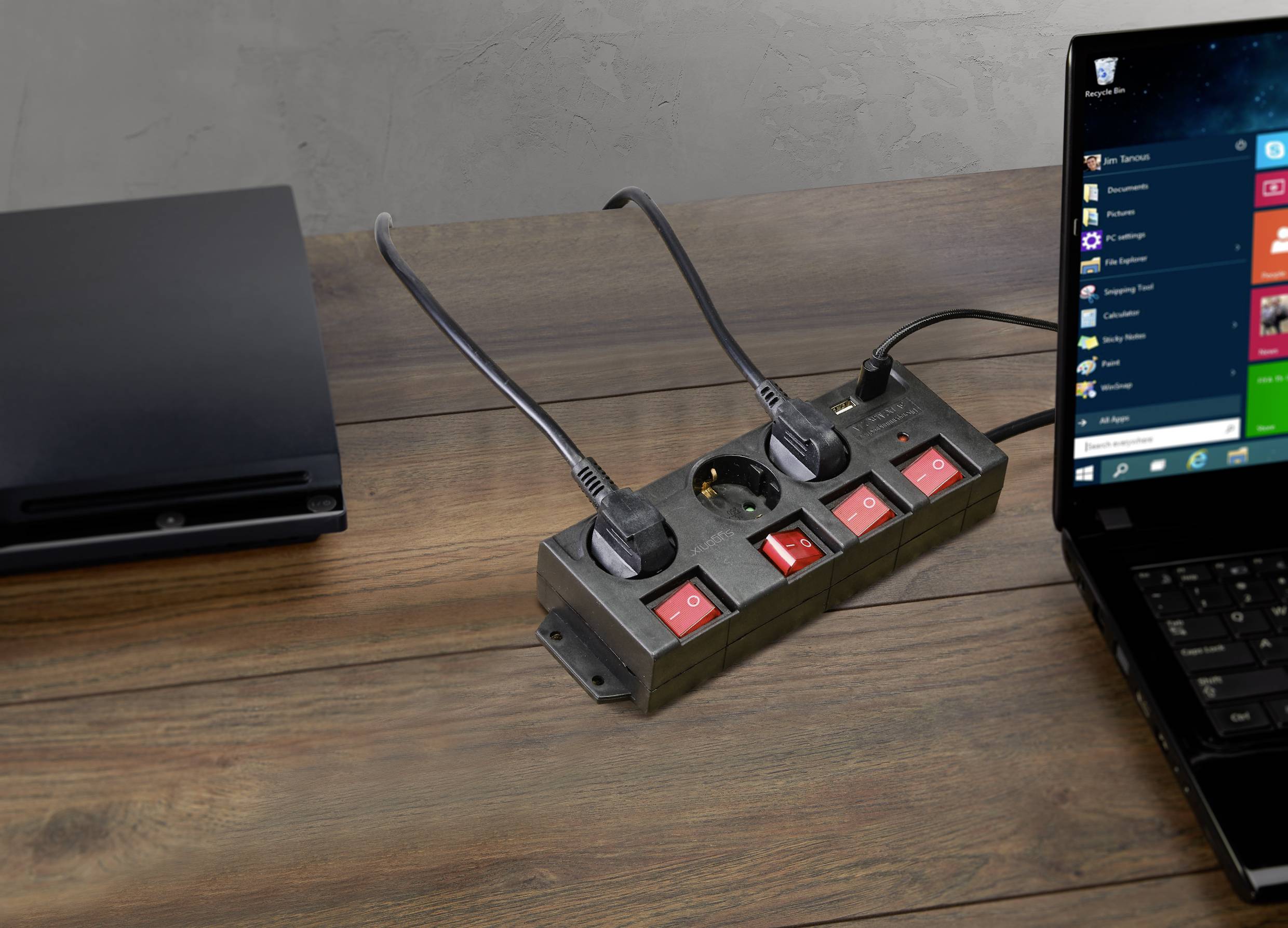 A laptop and a games console are connected to a power strip on a wooden table.