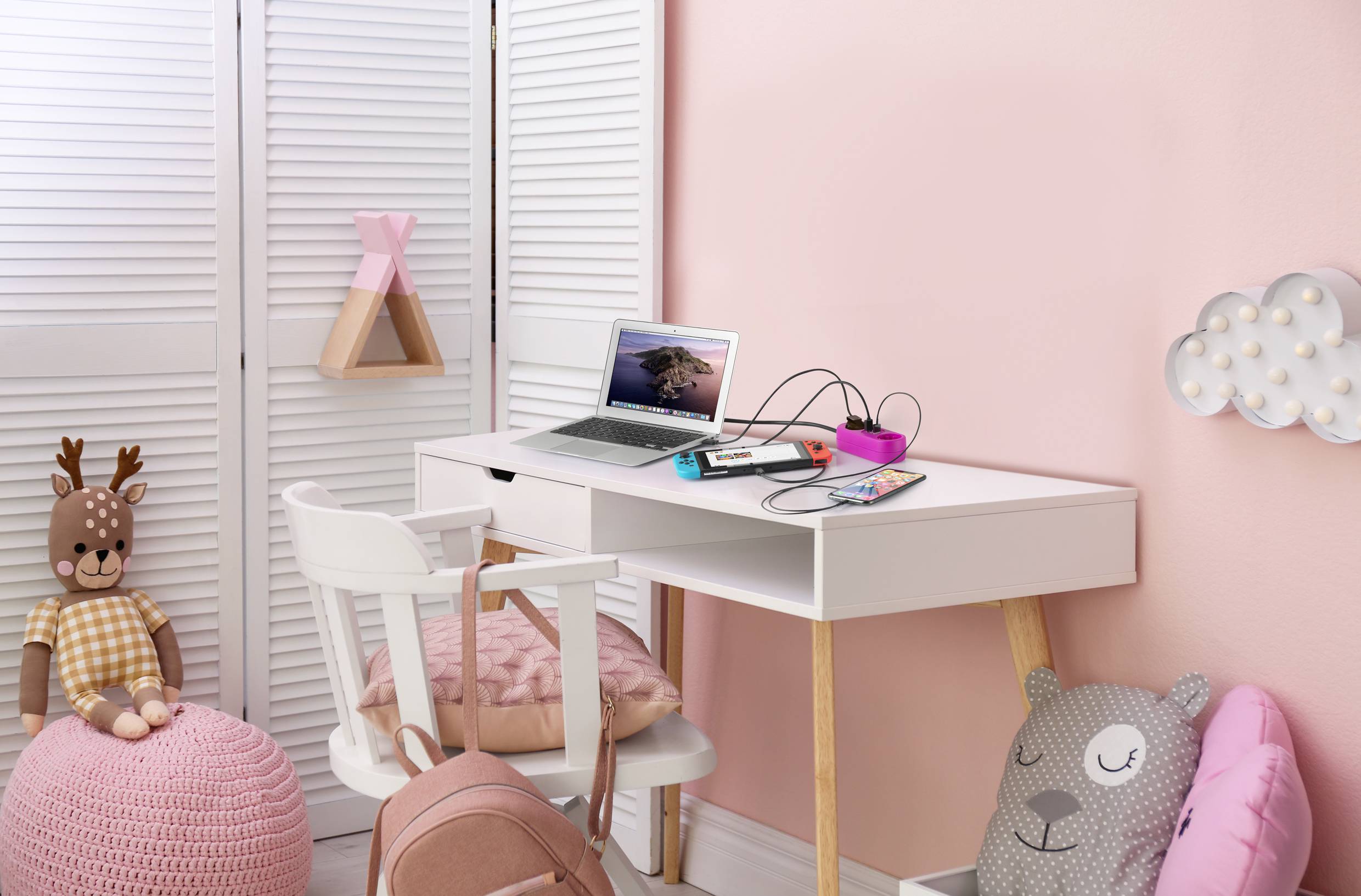 A children's bedroom with a desk, laptop, notebook and pens. Pink walls and decorations, including a cuddly toy and cushions.