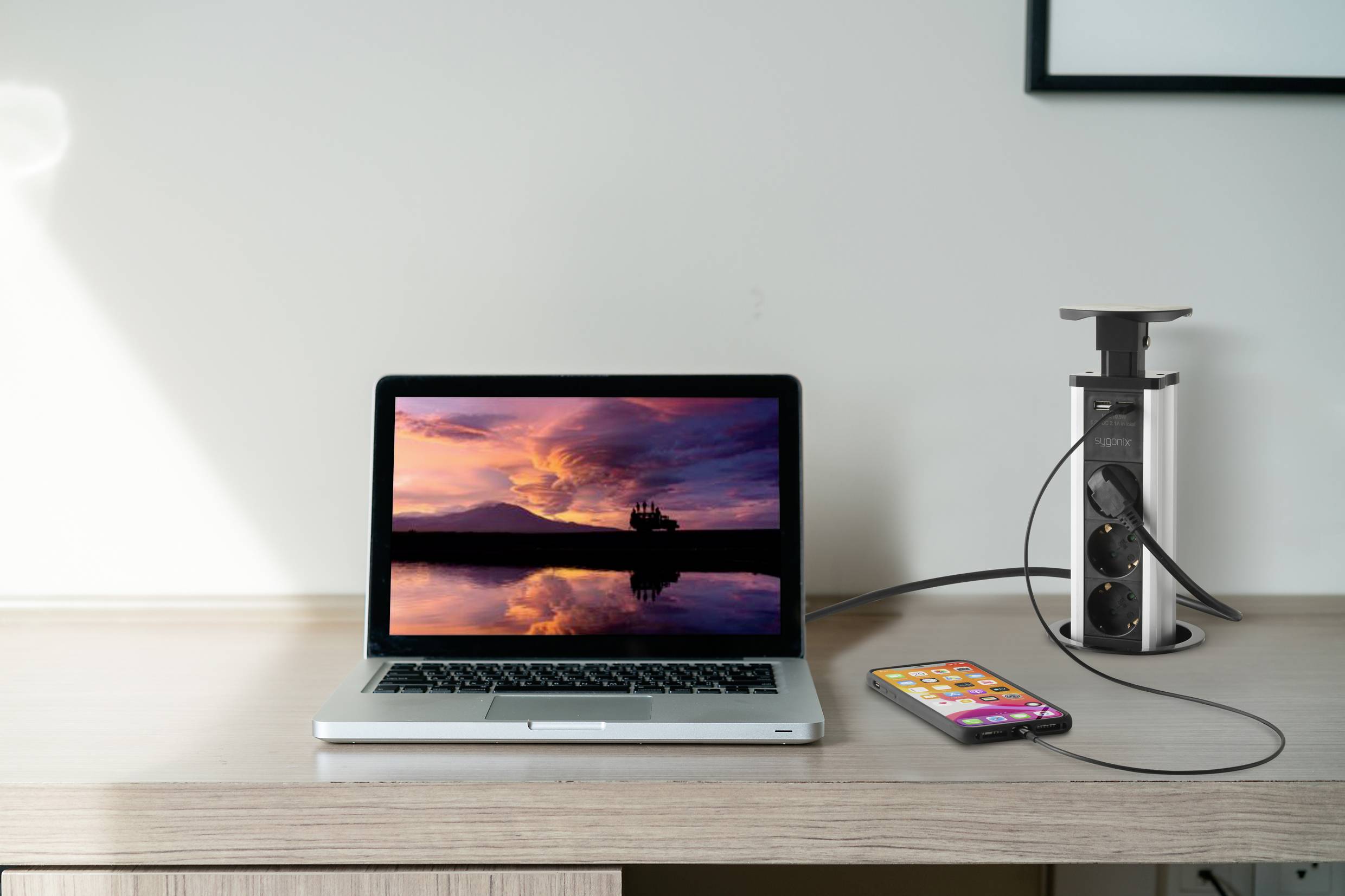 A laptop with a sunset screen and a smartphone are being charged on a table, connected to a pop-up power socket.