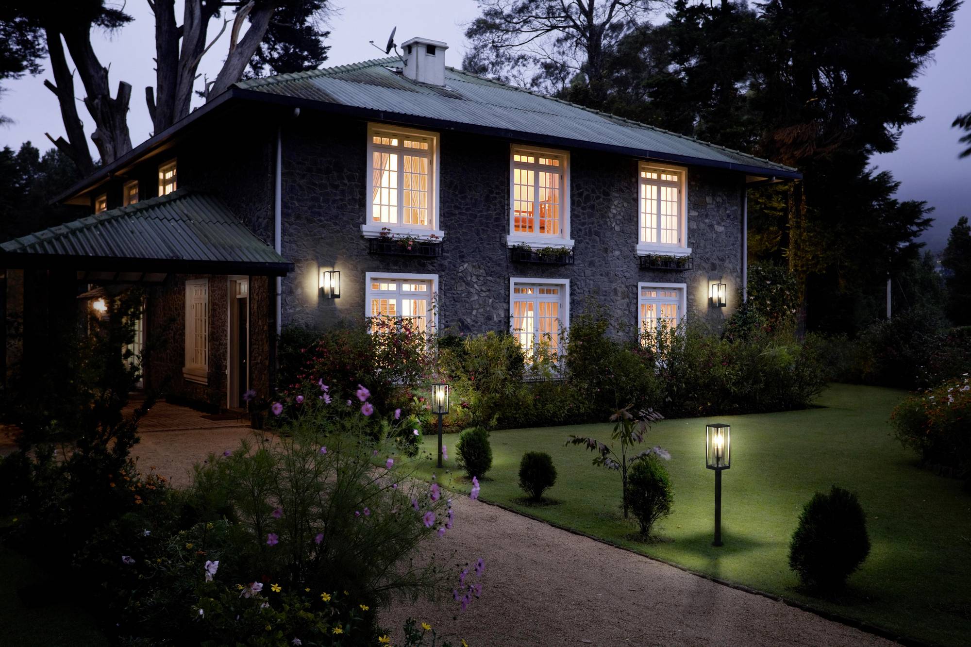 A two-storey house with illuminated windows in a garden. Dusk, with a lawn and flower beds surrounding the lit pathway.