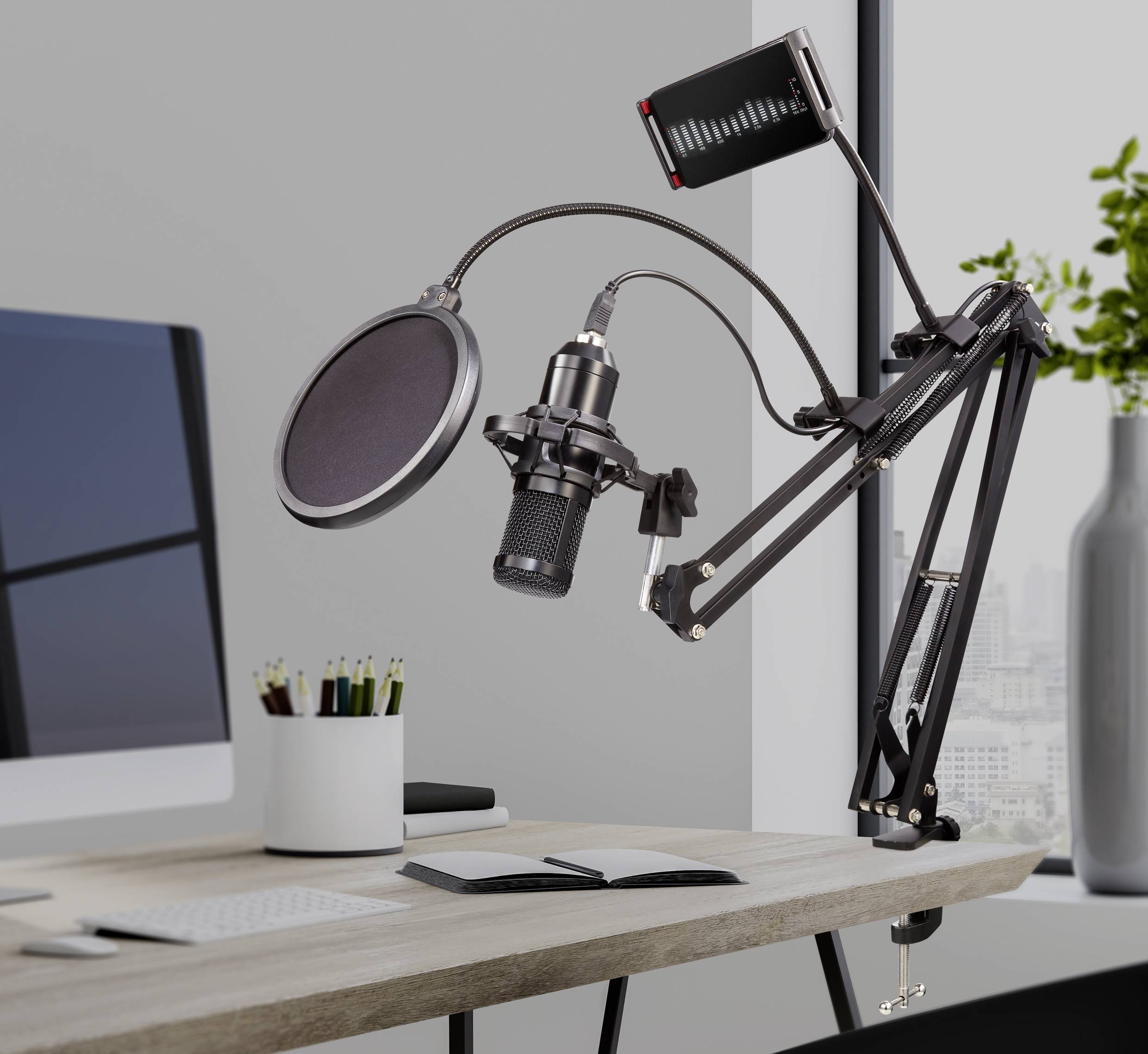 Microphone with pop shield on a boom arm, next to a monitor and notebooks on a desk, for podcast recording.