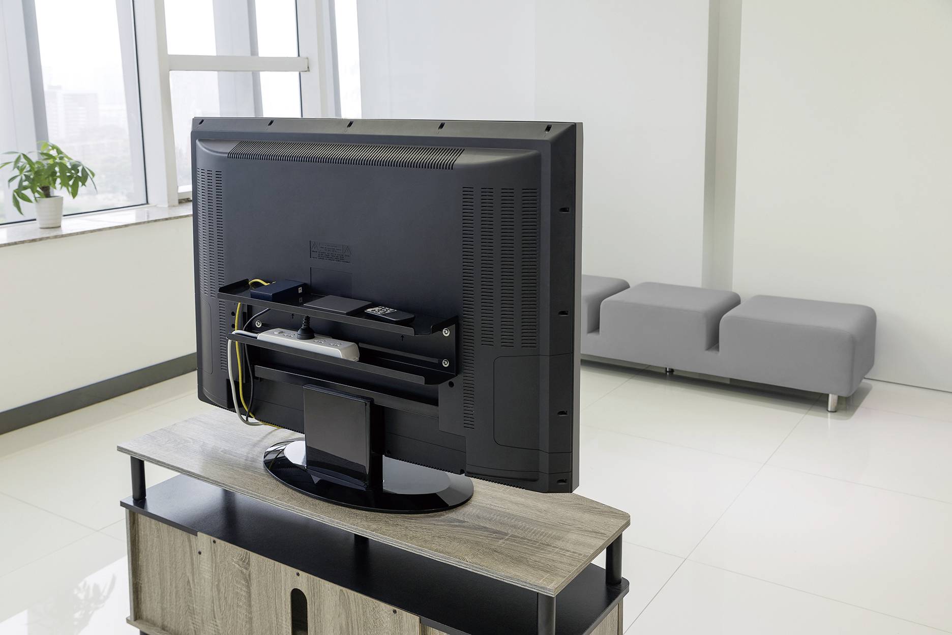 A flat-screen television is positioned at the back of a wooden piece of furniture in a bright room with grey seating and a plant by the window.