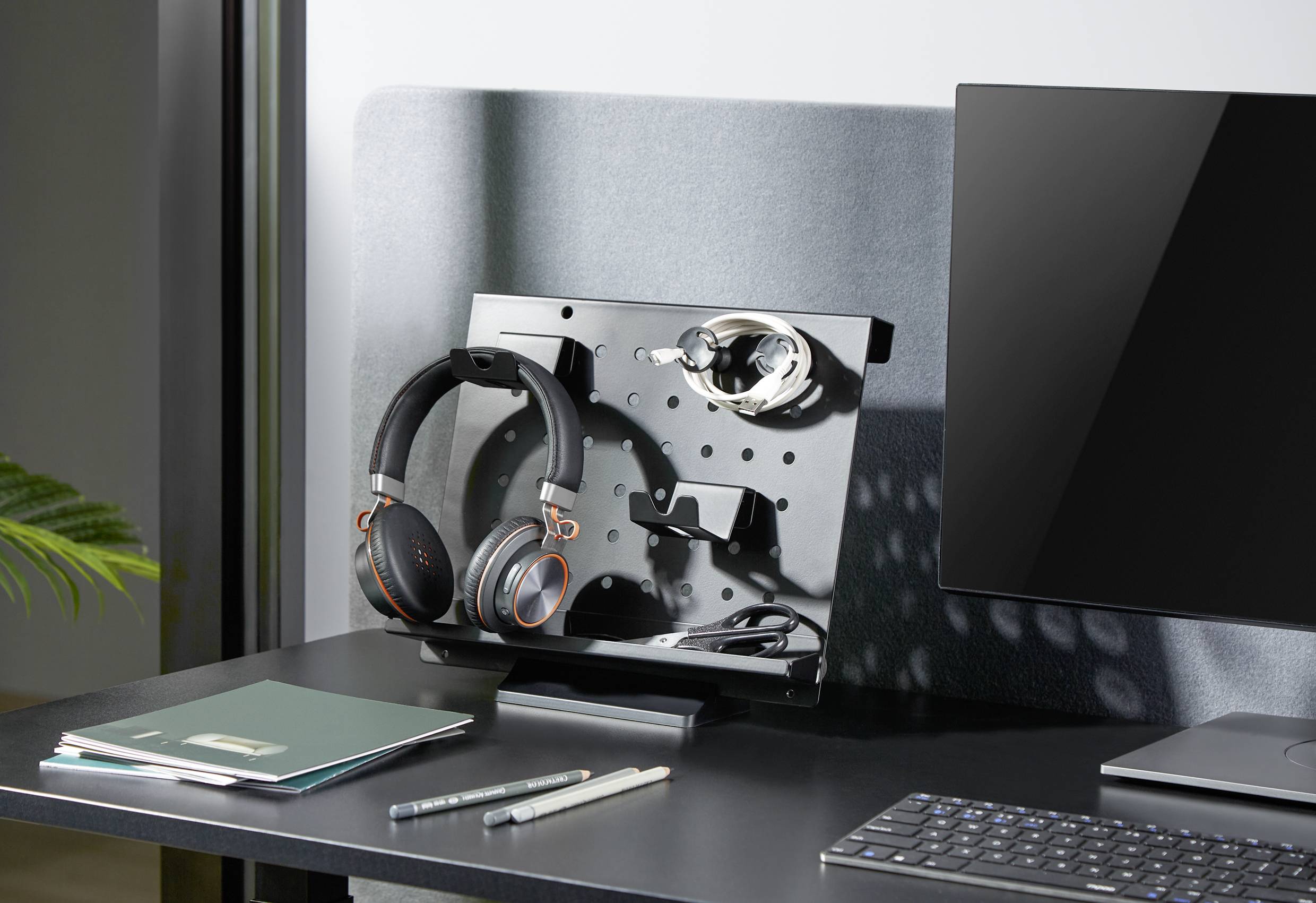 A desk with a computer, notebook, pens and a stand holding headphones and cables, with a monitor beside it.