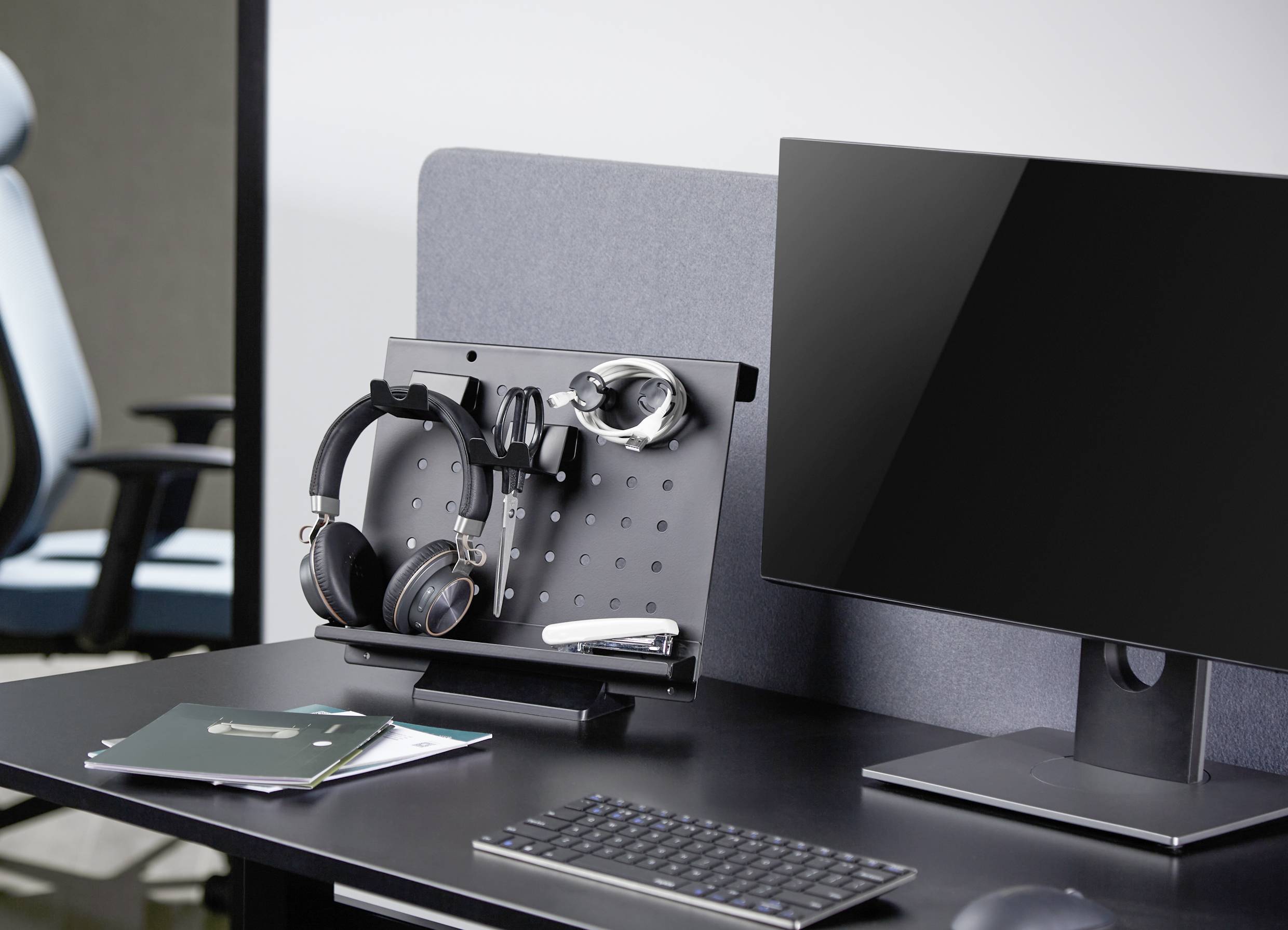 Desk with monitor, keyboard and organisational panel for headphones and office supplies. In the background, an empty chair.