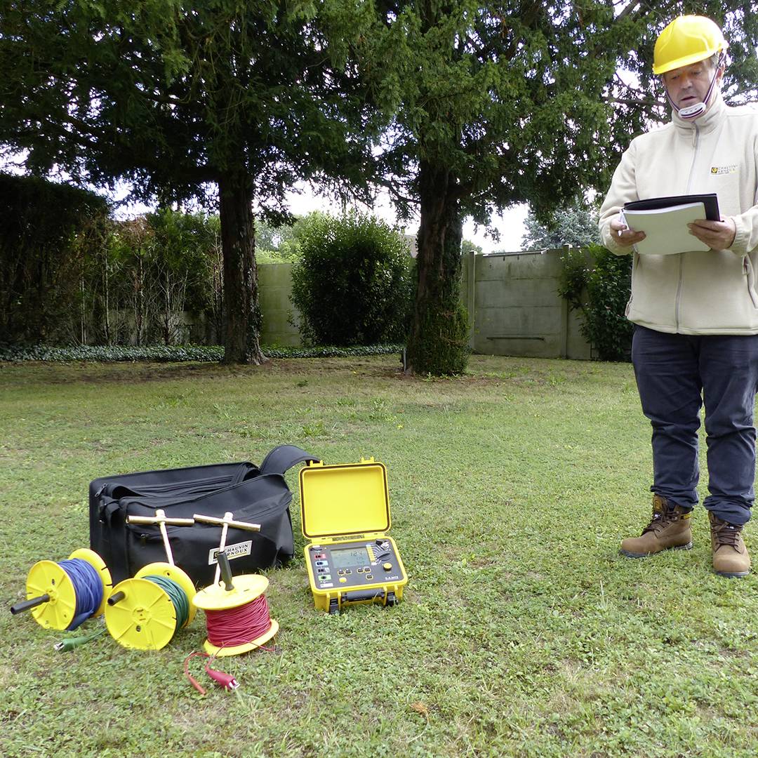 A worker wearing a hard hat inspects a measuring device outdoors. Cable reels and an open equipment housing lie beside him.