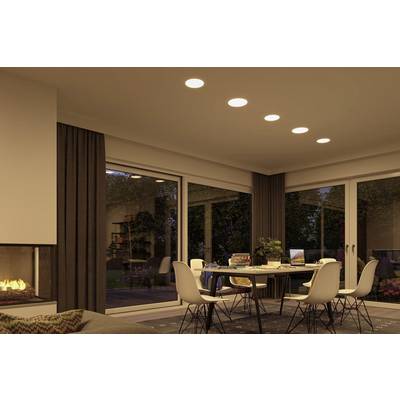 A modern living room with a dining table and six white chairs. Large windows offer a view of a garden. Ceiling lights are switched on.