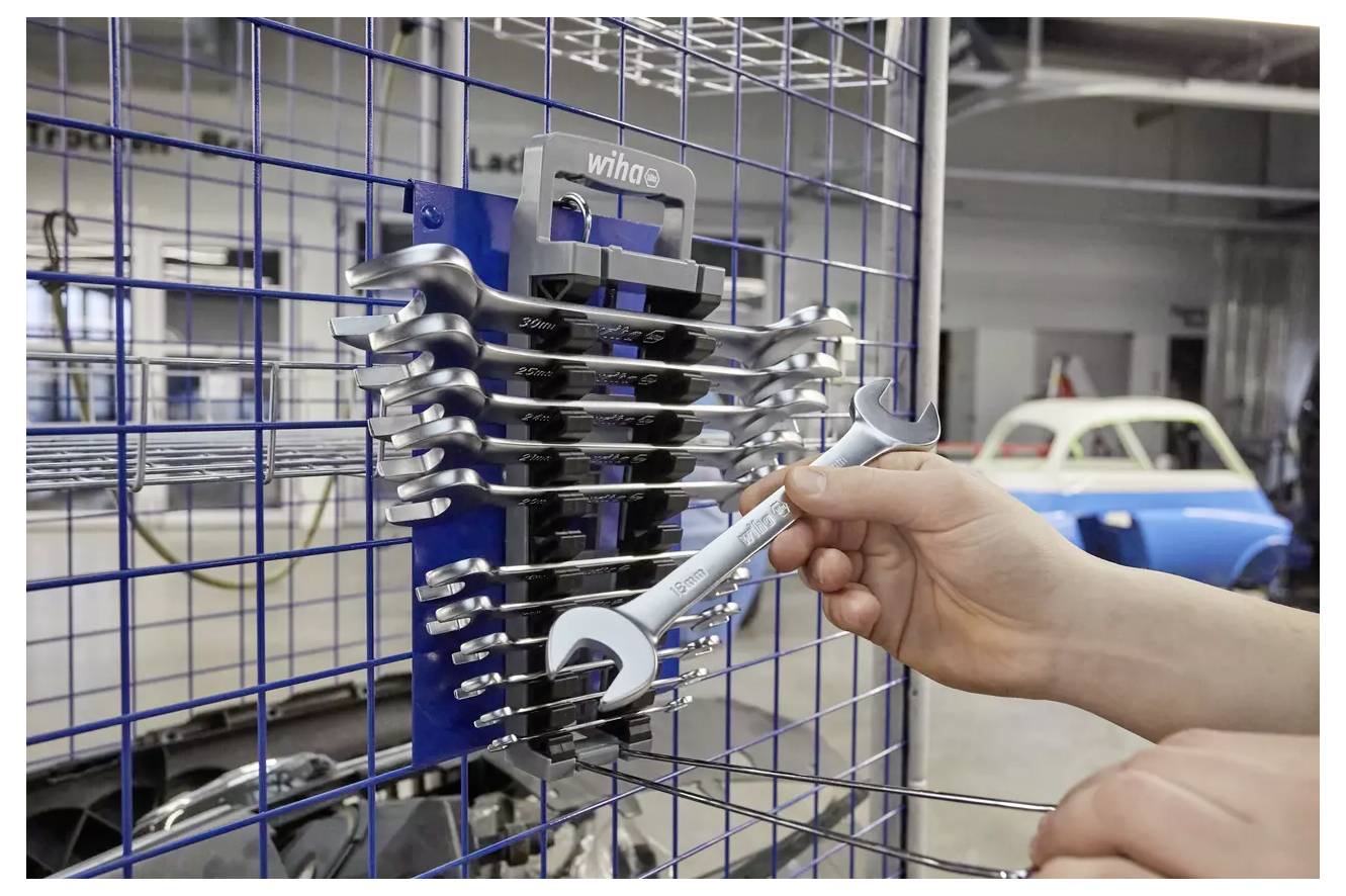A hand takes a wrench from a set organized on a metal grid in a workshop, with cars visible in the background.