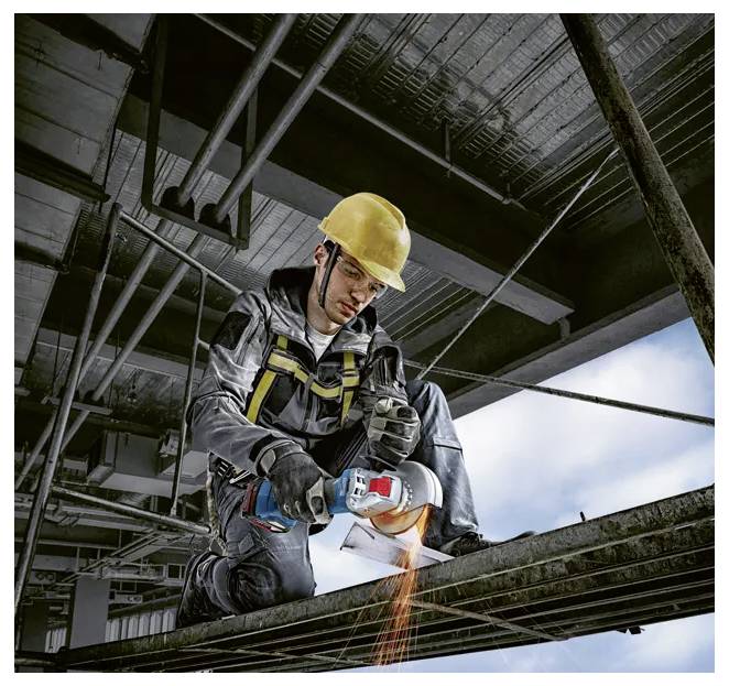 A construction worker wearing safety gear uses a power tool to cut metal beams while kneeling on a high-rise framework.