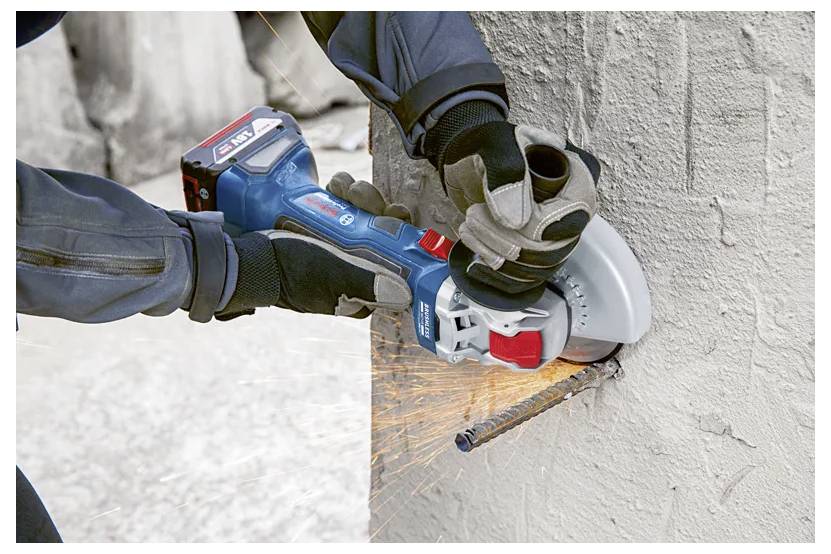 Person using a cordless angle grinder to cut a protruding metal rod from a concrete wall, with sparks flying from the cut.