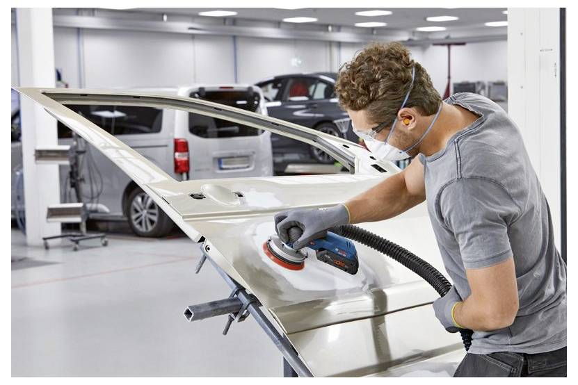 A person wearing safety gear is polishing a car door in an auto workshop. Several vehicles are visible in the background.