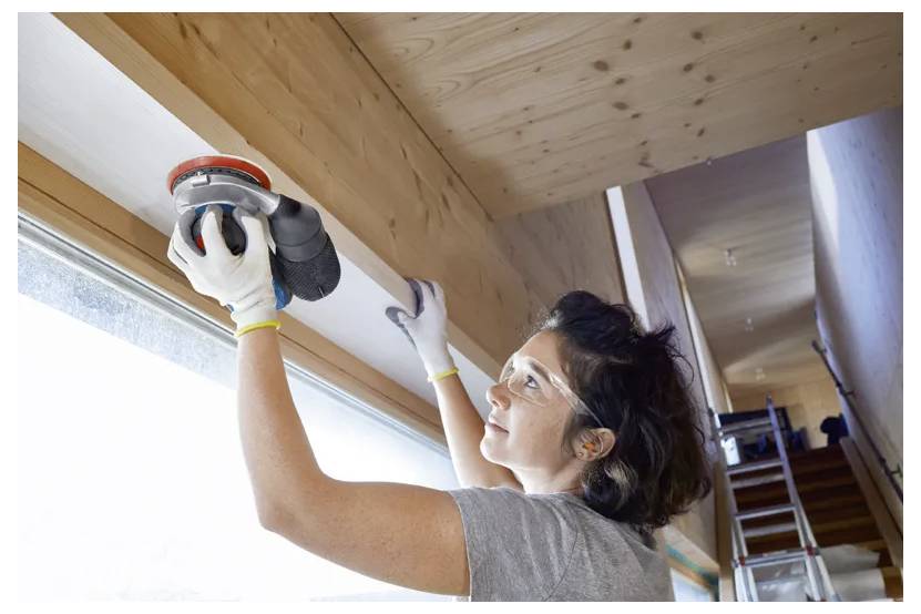 A woman wearing safety goggles sands a wooden beam indoors, focusing intently. A ladder and a large window are visible in the background, providing natural light.