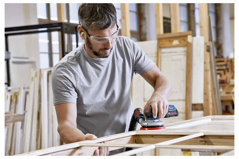 A person wearing safety goggles and ear protection uses an electric sander on a wooden frame in a workshop setting.