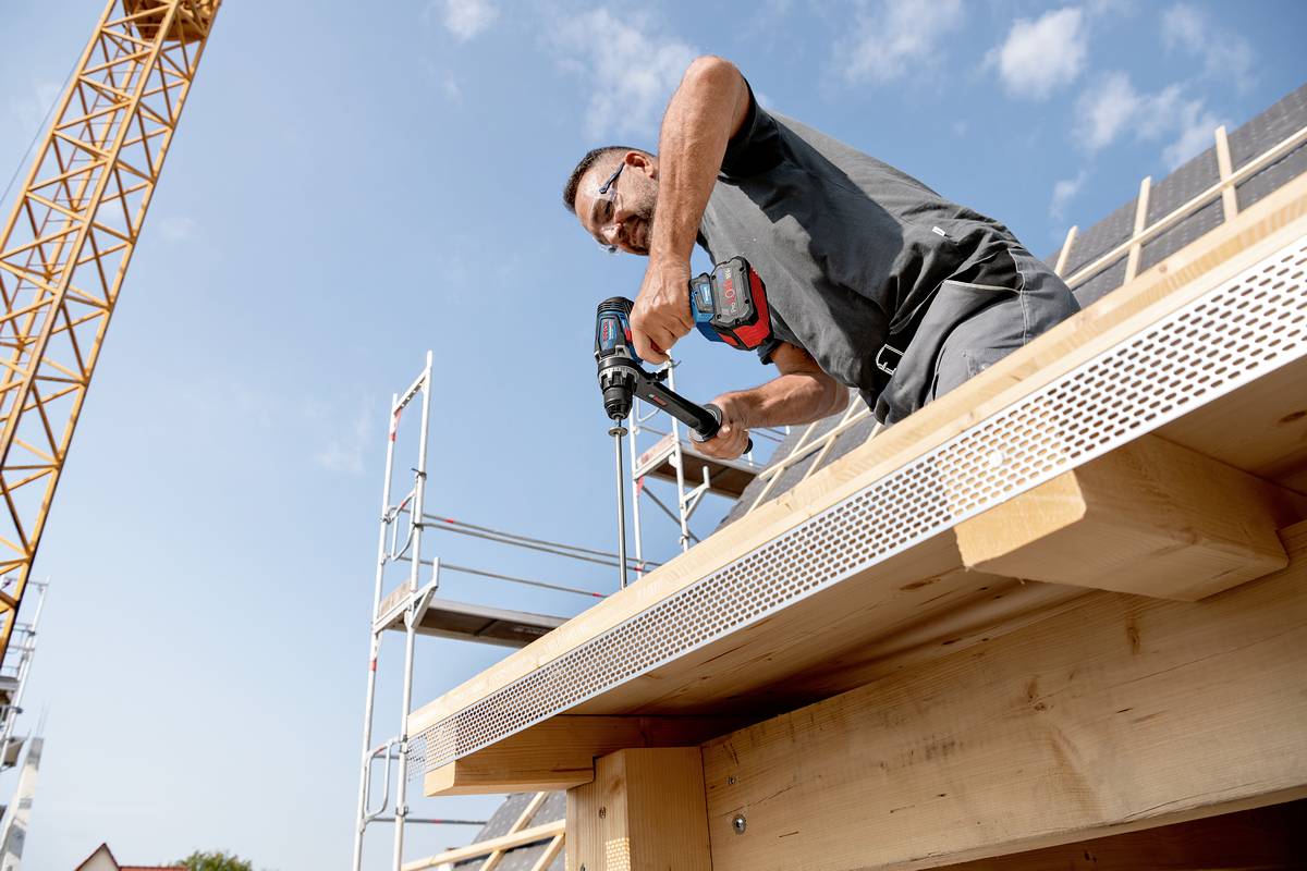 Worker on a construction site operating a power drill on a wooden roof. A crane and scaffolding are visible in the background.