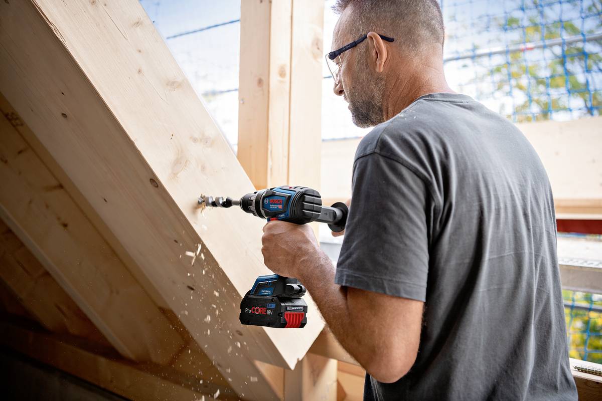 A man is using an electric drill to bore a hole into a wooden beam. A construction site is visible in the background.