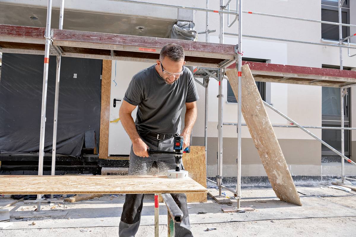 A man wearing safety glasses is cutting a wooden board with a saw on a construction site. Scaffolding is visible on the building in the background.