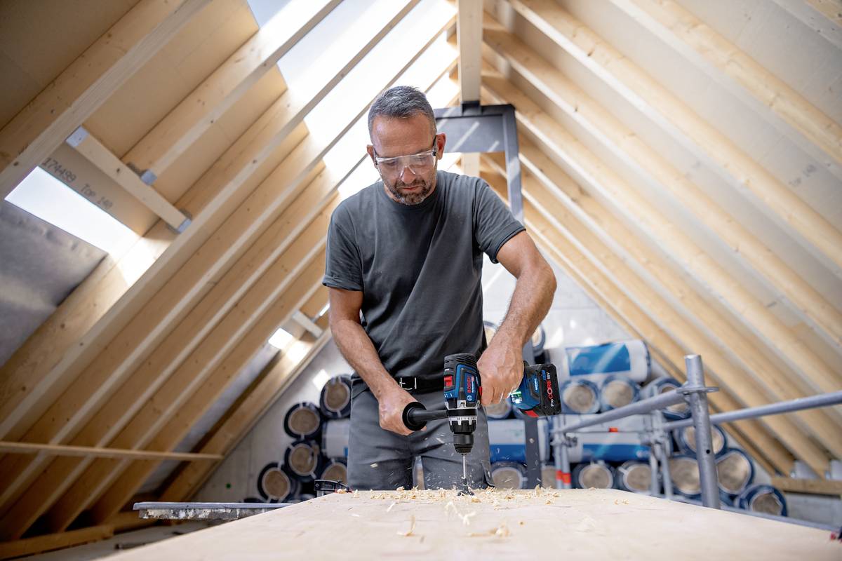A tradesman is drilling with a cordless drill in a wooden roof space. He is wearing safety glasses. The background shows construction site materials.