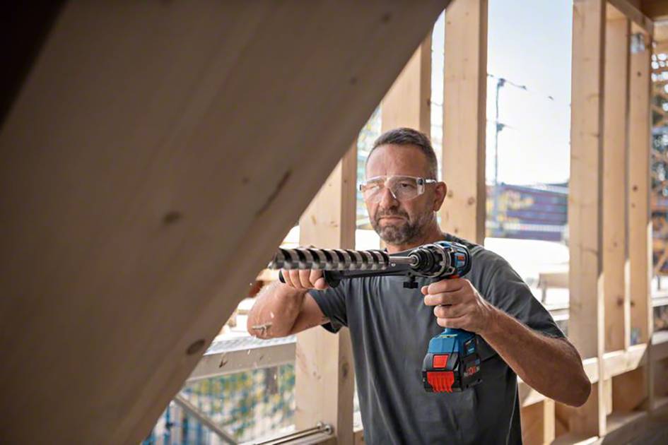 A man wearing safety glasses is working with a drill on a wooden frame at a construction site. Sunlight is filtering through the structure.