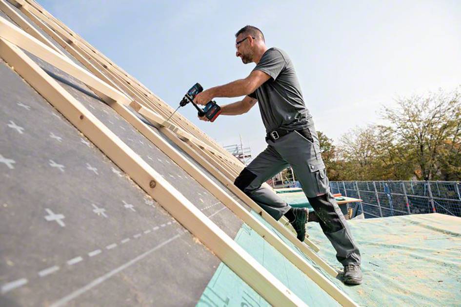 A man is working on a roof, drilling wooden battens with an electric tool. He is wearing protective clothing and scaffolding is visible in the surrounding area.