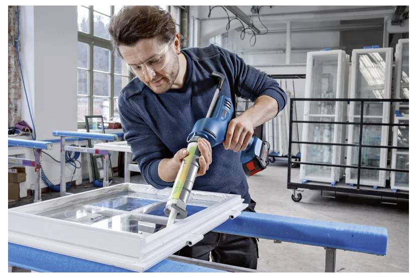 A person in safety glasses uses a power tool to seal a window frame in a workshop, surrounded by window panels and industrial equipment.