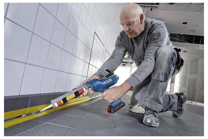 A person kneels on a tiled floor, using a blue electric caulking gun to seal the edge between the floor and wall tiles.
