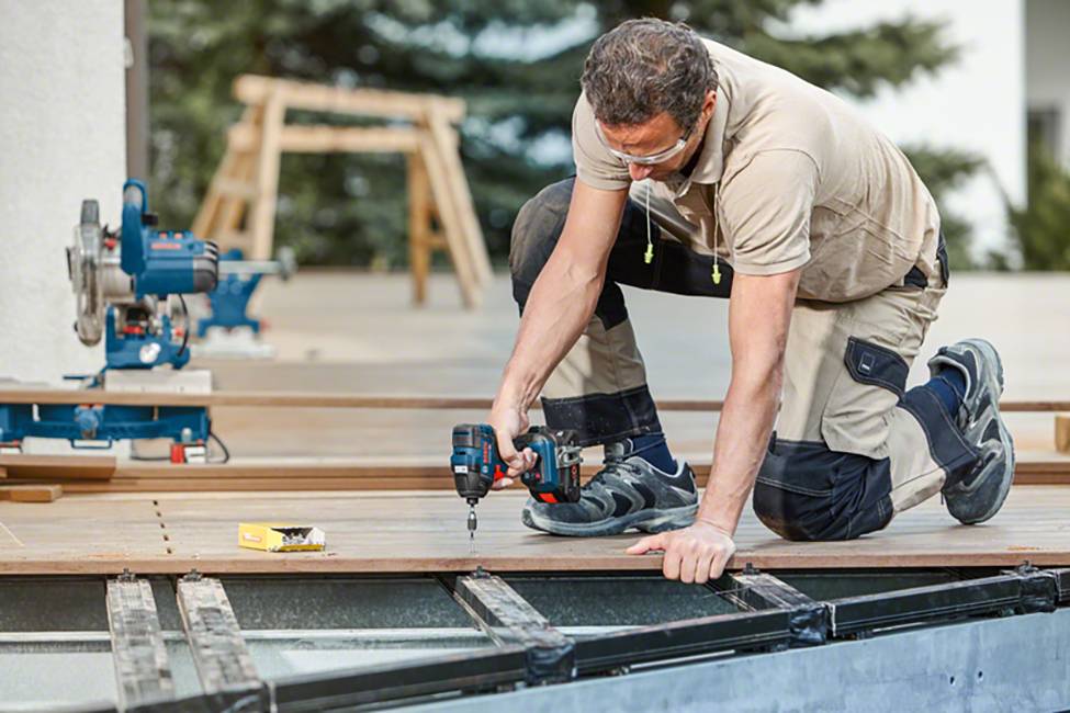 A man is kneeling and using a power drill to secure wooden floorboards. Tools and a ladder are visible in the background.