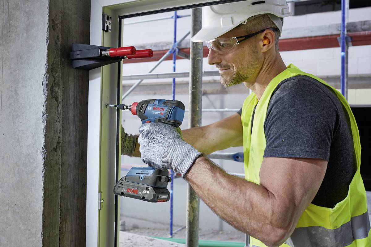 A construction worker in protective clothing is installing a window frame on a building site using a drill. He is wearing a hard hat and gloves.