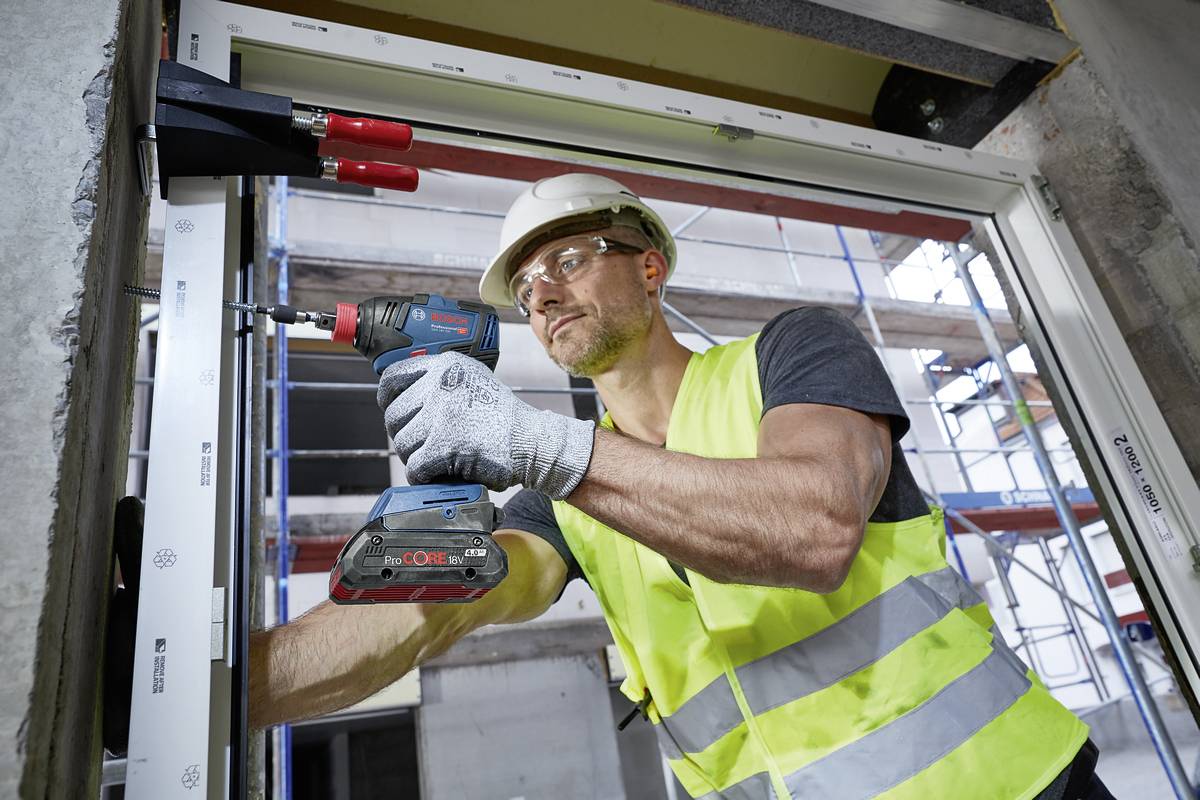 A construction worker wearing a hard hat and high-visibility vest is using a drill to work on a window opening in a building. A scaffolding structure is visible in the background.