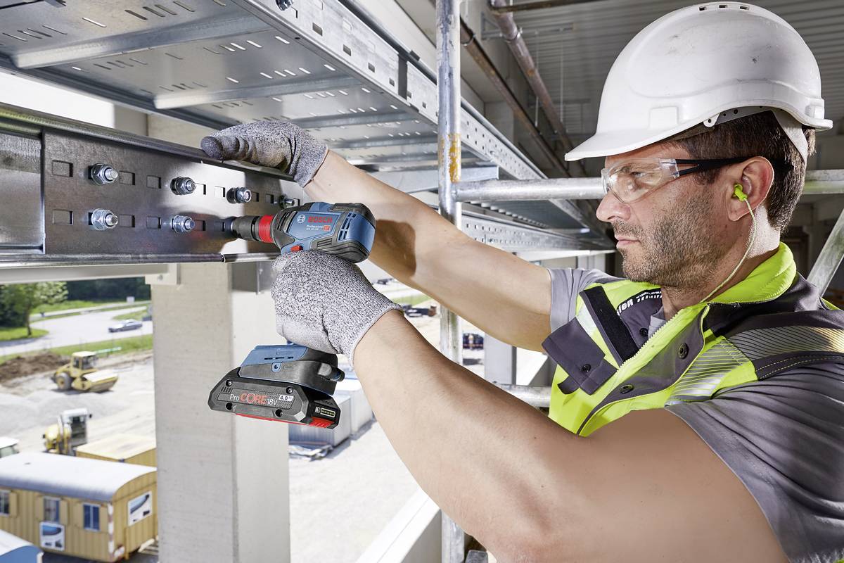 A construction worker wearing a hard hat is drilling metal on scaffolding at a building site. Construction vehicles and buildings can be seen in the background.