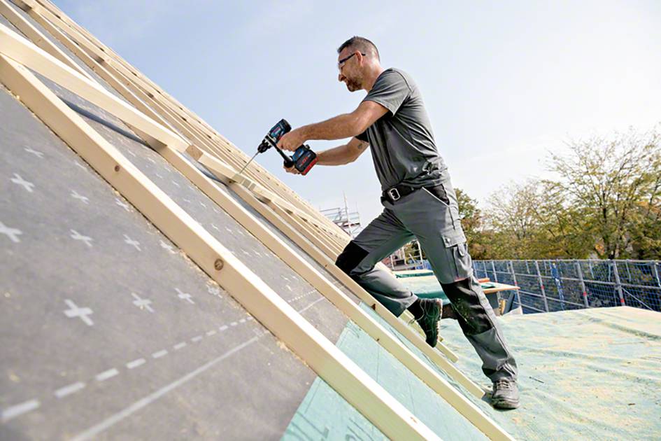 A man is working on a roof, using a drill to secure wooden battens. He is wearing work clothing and safety goggles.