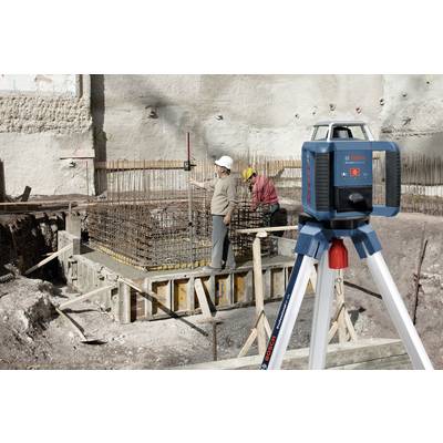 Construction workers are using levelling equipment on a building site; they are measuring the height of a concrete foundation fastening. In the foreground, a blue-grey laser device is mounted on a white tripod.
