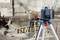Construction workers are using levelling equipment on a building site; they are measuring the height of a concrete foundation fastening. In the foreground, a blue-grey laser device is mounted on a white tripod.
