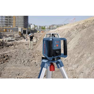 A construction worker is measuring a site using a rotating laser on a tripod. A construction site can be seen in the background.