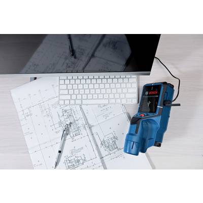 A laser rangefinder lies on a table with building plans, a keyboard and a monitor. A pen rests on the plans.