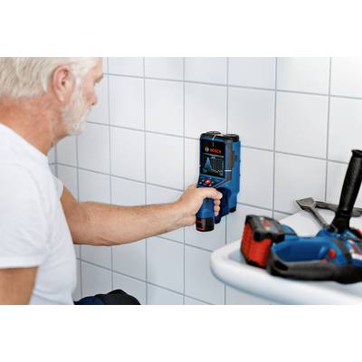 A man is using a Bosch distance measuring device on a tiled wall. Tools are lying on a small table beside him.