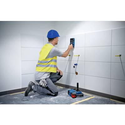 A construction worker is kneeling on the ground and measuring the moisture of a tiled wall with an electronic device. He is wearing a yellow hi-vis vest and a blue hard hat.