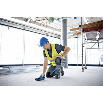 A construction worker wearing a hard hat and high-visibility vest is kneeling on a building site and using a measuring instrument.