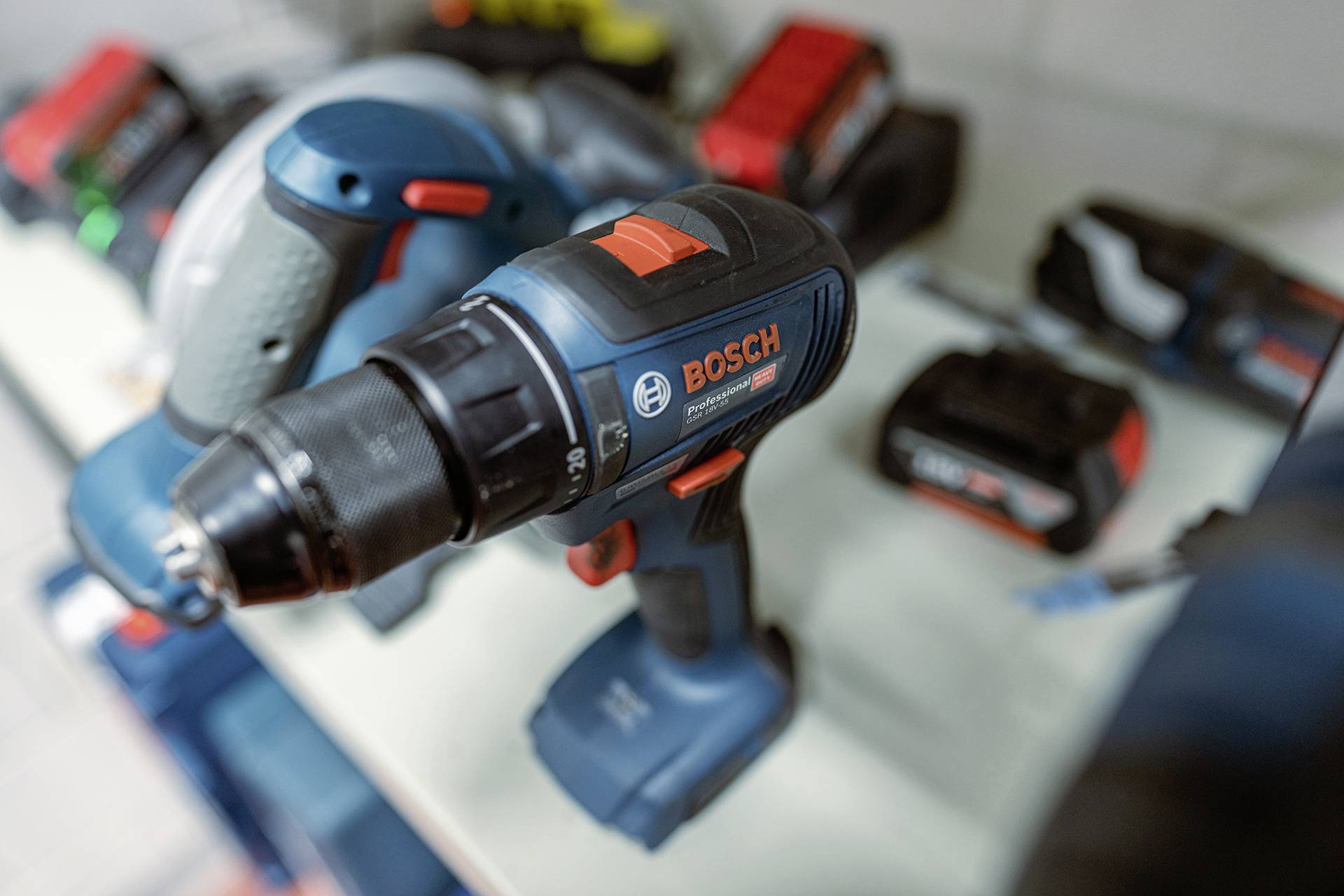 Cordless drill and accessories on a table. Additional tools blurred in the background. Dominant colour blue with red accents.
