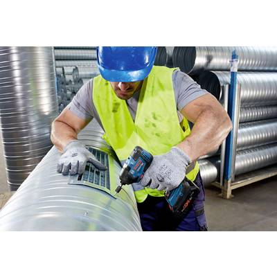 A worker wearing a blue hard hat and yellow hi-vis vest is using a cordless drill on a large ventilation pipe in a workshop.