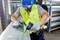 A worker wearing a blue hard hat and yellow hi-vis vest is using a cordless drill on a large ventilation pipe in a workshop.