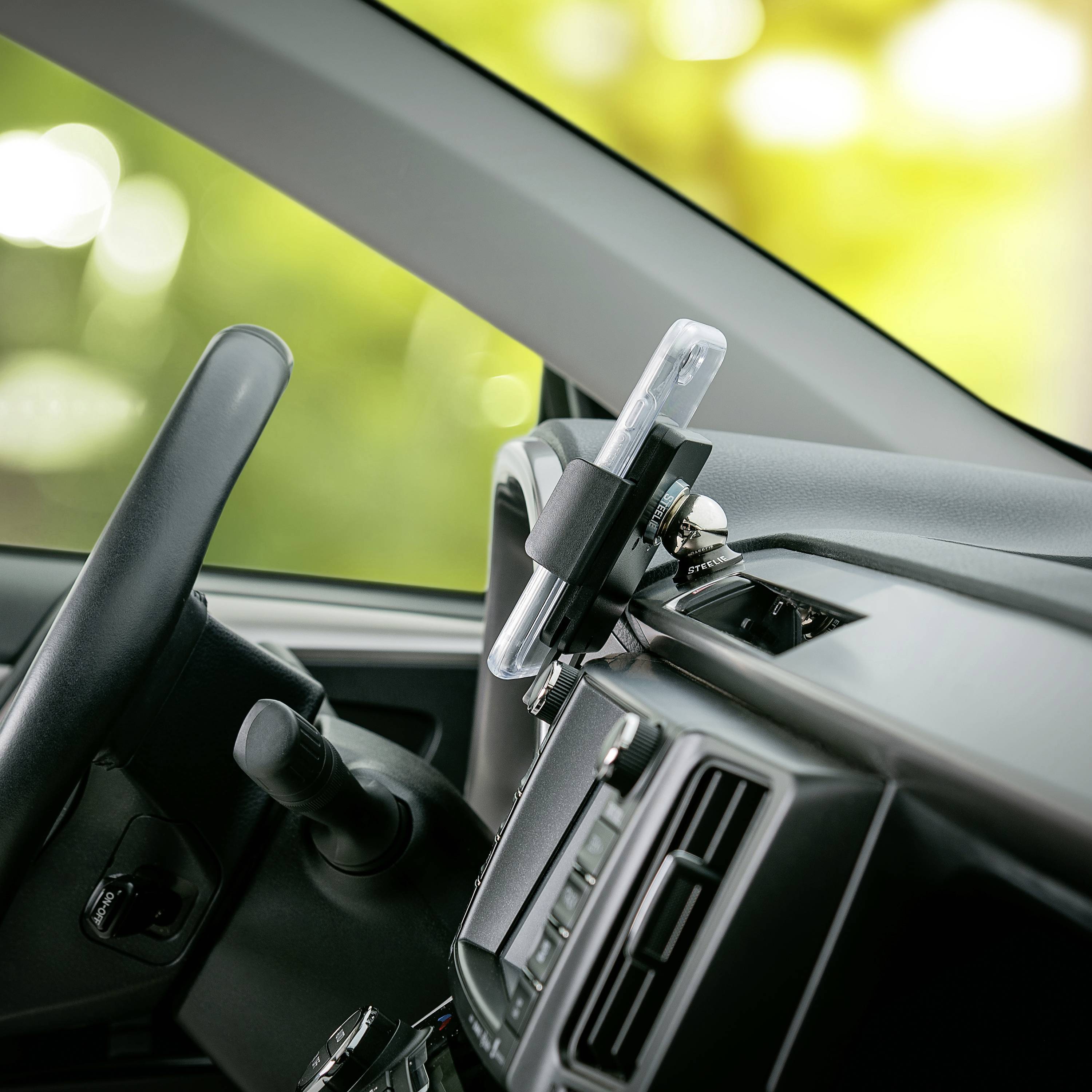 A smartphone in a mount in the car. It is inserted into the dashboard, close to the steering wheel. Sunlight is shining through the windows.