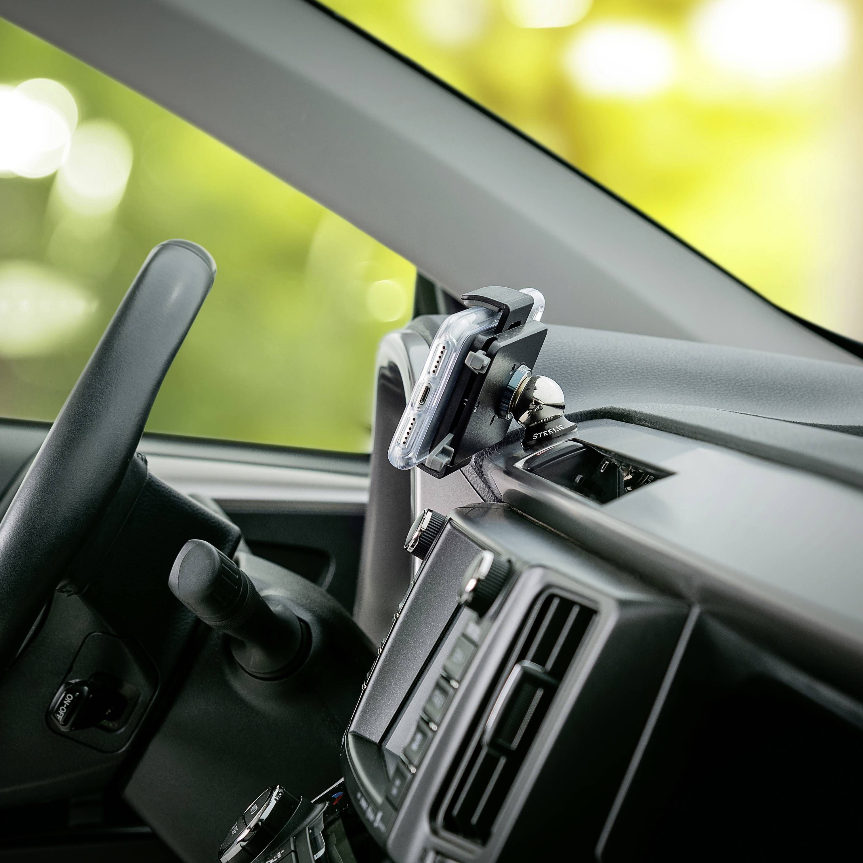 A mobile phone is mounted in a holder on a car's dashboard. The steering wheel and part of the dashboard are visible.