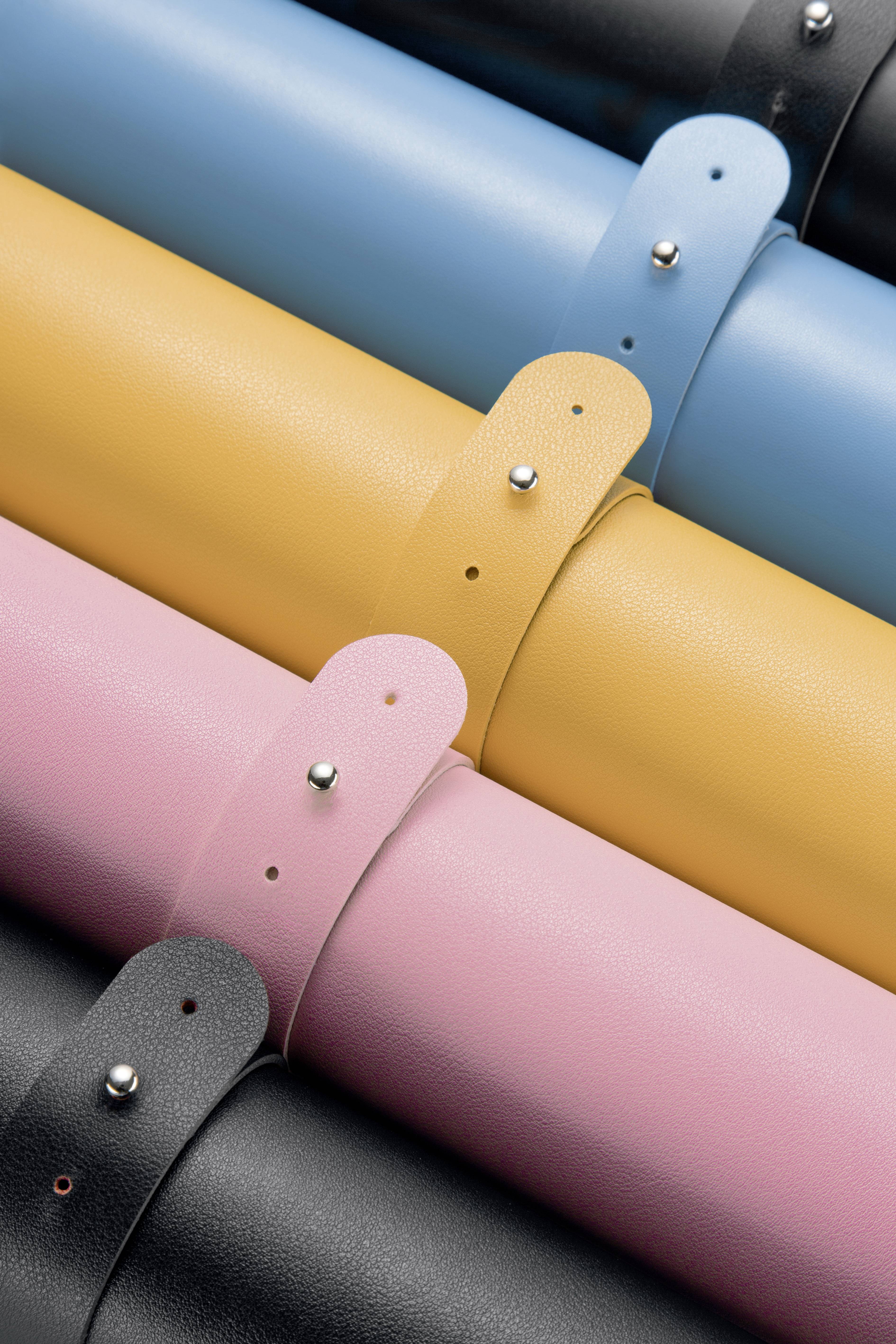 'Close-up of coloured faux leather rolls in blue, yellow, pink and black, stacked on top of each other, with attached straps and rivets.'