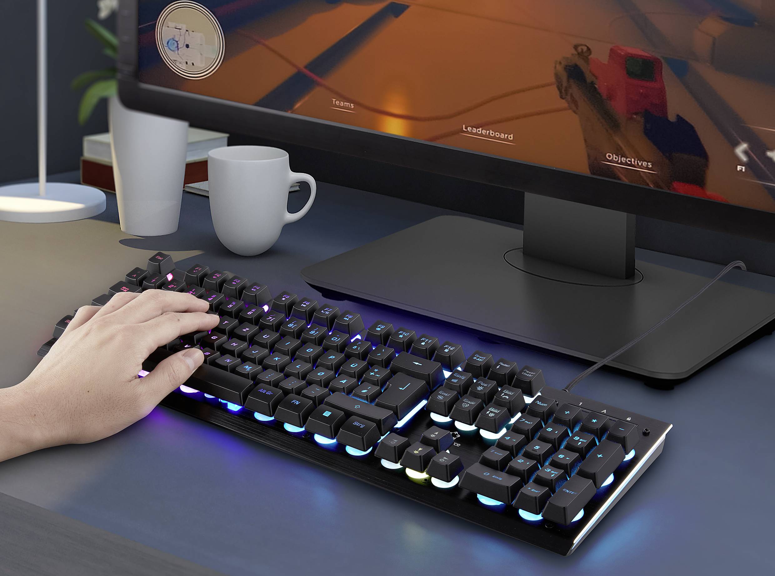 A person is typing on an illuminated gaming keyboard in front of a monitor. Two mugs are sitting on the table beside them.