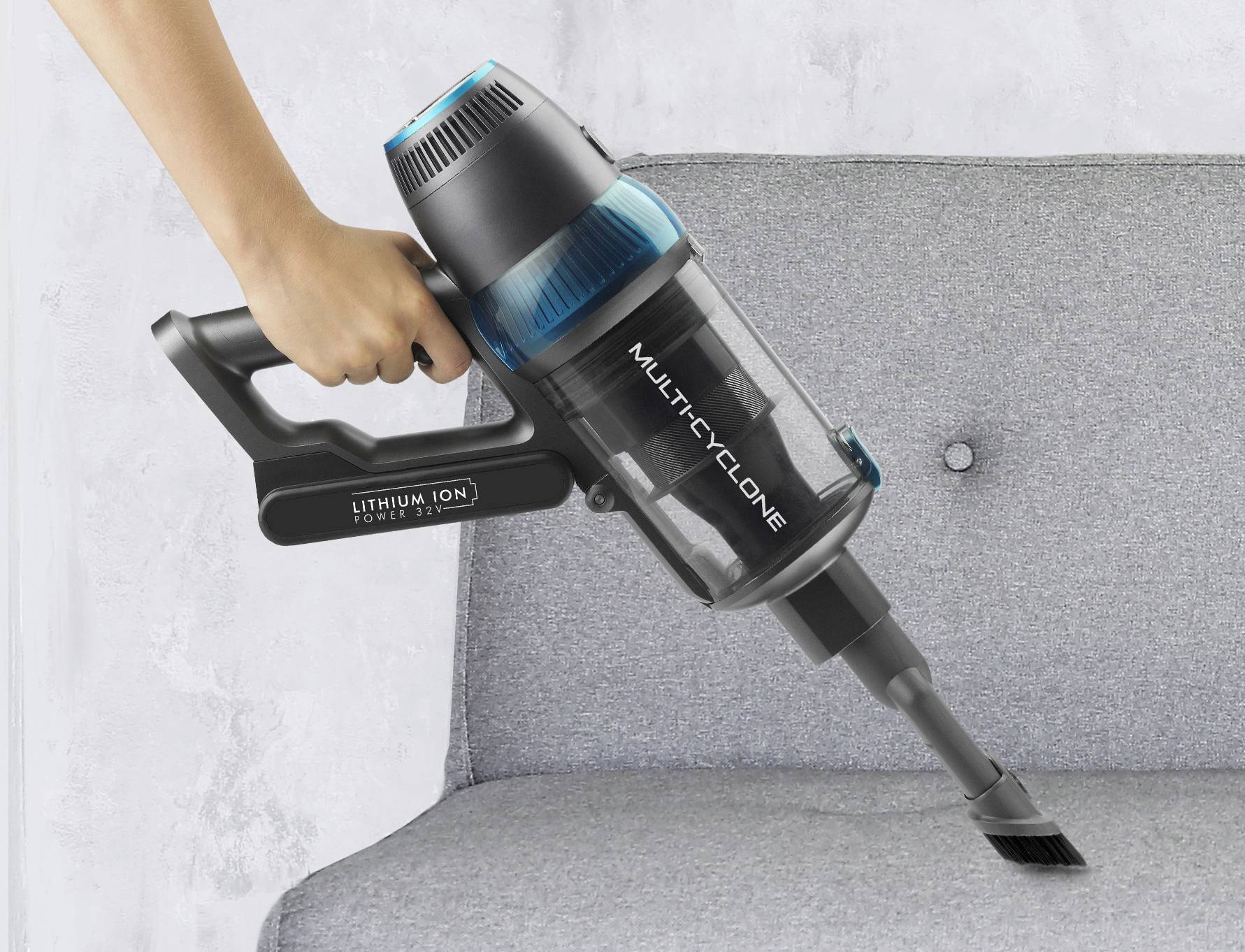 A person is holding a cordless handheld vacuum cleaner and cleaning a grey sofa. The vacuum cleaner has a blue accent colour and is marked with 'Multicyclone'.