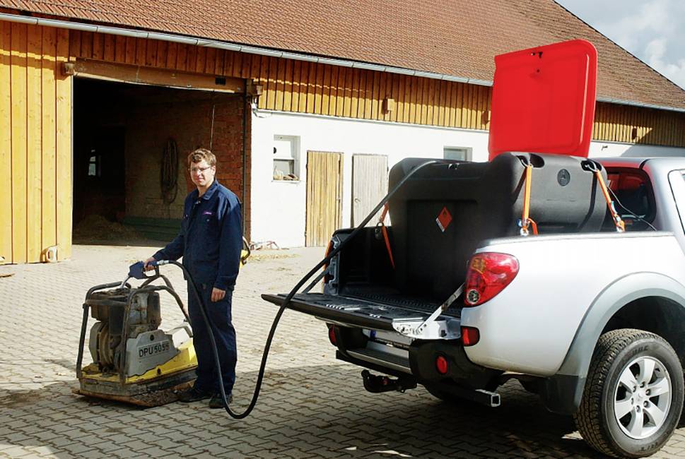 A person is refuelling a piece of construction machinery using a mobile fuel station mounted on a pickup truck, in front of a barn.